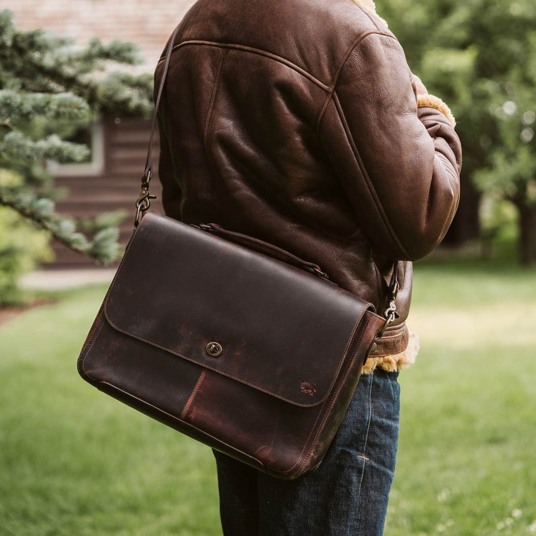 Man carrying Roosevelt Full-Grain Leather Messenger Bag in Dark Oak — back zipper pocket, worn crossbody in garden setting